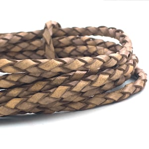 May include: Braided leather cord in a light brown color. The cord is tightly woven, creating a textured surface. The image shows several loops of the cord against a white background. This cord is suitable for jewelry making and crafting.