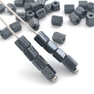 Może przedstawiać: Zbliżenie ciemnoszarych, sześciokątnych koralików nawleczonych na srebrny drut. Koraliki mają metaliczny połysk i są ułożone w rzędach, z kilkoma luźnymi koralikami w tle. Koraliki do tworzenia biżuterii.