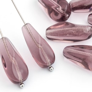 Puede incluir: Un grupo de ocho cuentas de vidrio en forma de lágrima de color morado claro con motas plateadas. Las cuentas son lisas y tienen un acabado brillante.