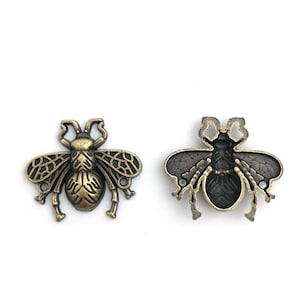 Può includere: Due ciondoli a forma di ape in oro antico, uno con un designo dettagliato e l'altro con un centro vuoto.