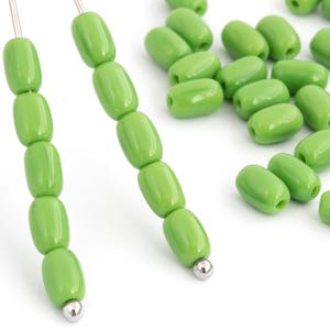 Op de afbeelding: Een streng groene glazen kralen op een zilveren draad. De kralen zijn ovaalvormig en hebben een glanzende afwerking. Er zijn ook losse groene glazen kralen verspreid rond de streng.