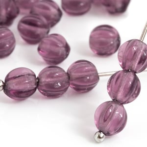 Puede incluir: Primer plano de varias cuentas de vidrio púrpura translúcidas con una textura acanalada. Algunas cuentas están ensartadas en un alambre plateado, mientras que otras están dispersas. Las cuentas tienen forma esférica y un acabado brillante.