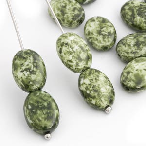 May include: Oval-shaped green and white beads with a speckled pattern. The beads are strung on silver-colored wire, with a silver bead at the end of each strand. The beads are smooth and glossy, and are arranged on a white surface.