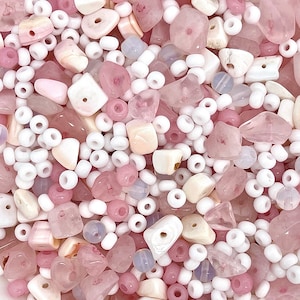 Pode incluir: Uma mistura de contas e pedras rosa e brancas. A variedade inclui várias formas e tamanhos, como pequenas contas brancas, pedras arredondadas e pedras rosa maiores e irregulares. A paleta de cores é suave e feminina.