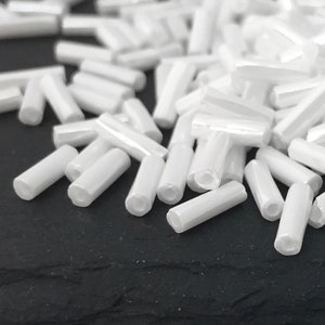 Op de afbeelding: Een stapel witte glazen zaadkraaltjes, ook wel bugle-kraaltjes genoemd, op een zwart oppervlak. De kralen zijn cilindrisch en hebben een gladde, glanzende afwerking.