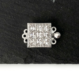 May include: A square, silver-coloured jewellery clasp with a grid of small, clear stones set in the centre. The clasp has decorative loops on each side and a small, round closure mechanism. The piece is displayed on a dark, textured surface.