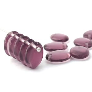 Puede incluir: Cuentas de vidrio ovaladas y translúcidas de color púrpura. Algunas cuentas están ensartadas, otras dispersas. Las cuentas tienen una superficie lisa y pulida y un pequeño agujero para ensartar. Fondo blanco.