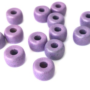 May include: A collection of small, cylindrical wooden beads in a muted purple hue. Each bead has a central hole, and the beads are scattered against a white background. These beads are suitable for jewelry making and craft projects.
