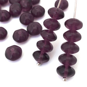 May include: Close-up of dark purple glass beads. Some beads are loose, while others are strung on wire. The beads have an oval or rounded shape with a faceted surface. A small silver bead is at the end of each strand.