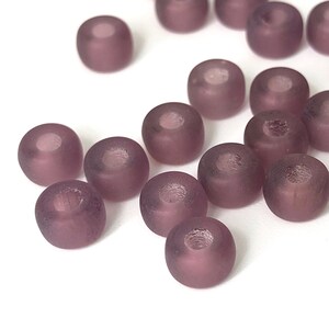May include: A collection of matte, round beads in a muted purple hue. Each bead has a central hole, and the surface appears slightly textured. These beads are suitable for jewelry making and crafting projects.