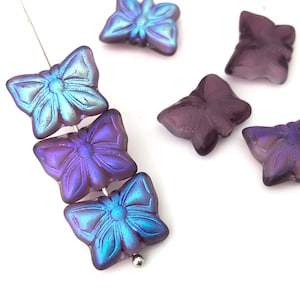 Puede incluir: Un grupo de cuentas de vidrio con forma de mariposa iridiscentes de color morado y azul. Las cuentas están enhebradas en un alambre plateado.