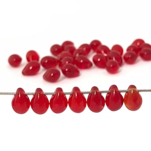 Könnte beinhalten: Rote, tropfenförmige Perlen, lose und auf einem Draht aufgereiht. Die Perlen sind von transluzenter, glänzender roter Farbe. Der Hintergrund ist weiß. Geeignet für die Schmuckherstellung.