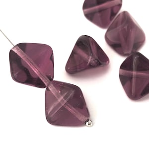 Puede incluir: Una hebra de cuentas de vidrio morado con forma cuadrada facetada. Las cuentas están enhebradas en un alambre plateado.