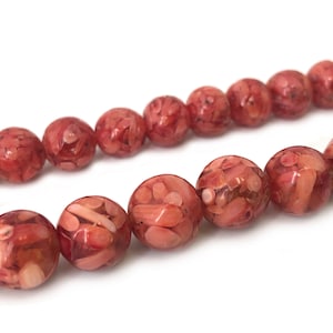 Puede incluir: Una hebra de cuentas de coral, cada cuenta es redonda, de color rojo y blanco con un patrón moteado.
