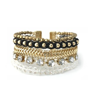 Könnte beinhalten: Ein mehrschichtiges Armband mit goldfarbenen Perlen, einer schwarzen Kordel, goldenen Kettengliedern, klaren Kristallen und klaren Perlen. Das Armband hat einen goldfarbenen Verschluss. Der Schmuck ist vor einem weißen Hintergrund platziert.