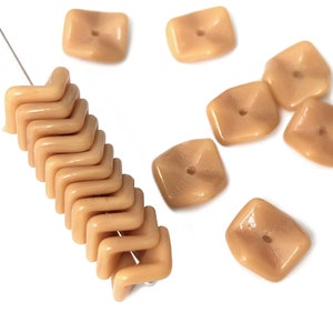 Può includere: Una collezione di perline di vetro beige, alcune sono quadrate con un foro al centro, e altre hanno una forma a zig-zag.