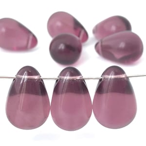 Puede incluir: Primer plano de varias cuentas translúcidas en forma de lágrima en tonos morados. Algunas cuentas están sueltas, mientras que otras están ensartadas en un alambre plateado. Las cuentas tienen un acabado liso y brillante sobre un fondo blanco.