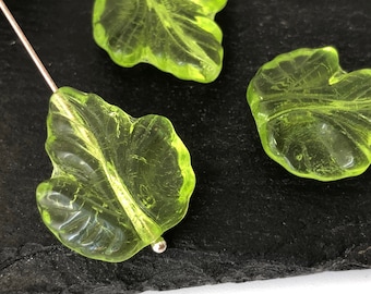 Op de afbeelding: Close-up van drie doorschijnende, limoengroene, bladvormige kralen. Elke kraal heeft gedetailleerde nerven en een licht getextureerd oppervlak. De kralen liggen op een donkere, getextureerde ondergrond.