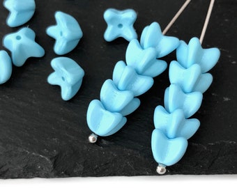 Puede incluir: Cuentas en forma de flor de color azul claro. Algunas están sueltas, mientras que otras están ensartadas con alfileres plateados. Las cuentas tienen un acabado mate y están dispuestas sobre una superficie gris oscuro. Estas cuentas son adecuadas para la fabricación de joyas.