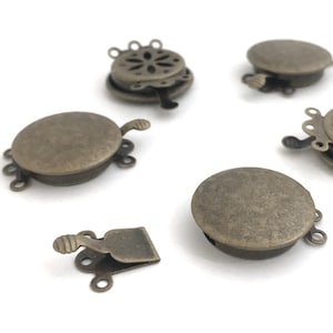 Könnte beinhalten: Fünf antike goldfarbene Metallverschlüsse zum Schmuckmachen. Jeder Verschluss hat eine runde, flache Basis mit einem dekorativen Design und drei Schlaufen zum Anbringen an eine Halskette oder ein Armband.