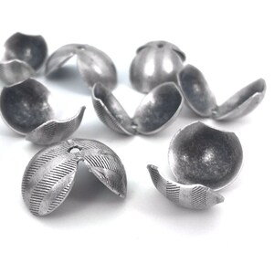 Puede incluir: Un grupo de cuentas de metal plateado con una superficie texturizada. Las cuentas están divididas en dos mitades y tienen un pequeño agujero en el centro.