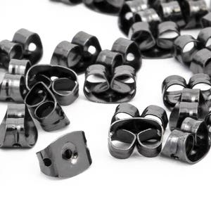 May include: A collection of black earring backs scattered on a white surface. The earring backs are made of metal and have a butterfly shape with a hole for the earring post.