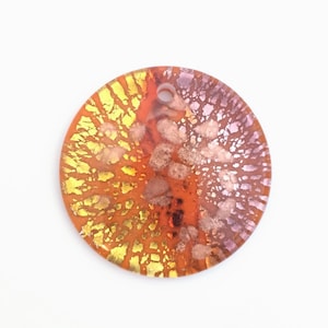 May include: A round, decorative pendant with a hole at the top. The pendant features a blend of colors, including yellow, orange, and purple, with a textured, abstract design. The pendant is set against a white background.