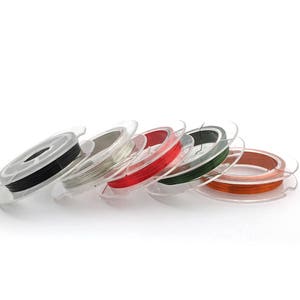 May include: Spools of colorful wire in various colors, including black, silver, red, green, and orange, are wound around clear plastic spools. The spools are arranged in a row on a white surface.