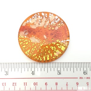 May include: A round, orange glass pendant with gold and white accents. The pendant has a diameter of approximately 2 inches. The design features a network of gold and white lines and shapes, creating a unique, decorative piece. The pendant is set against a white background.