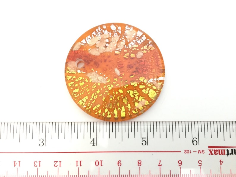 May include: A round, orange glass pendant with gold and white accents. The pendant has a diameter of approximately 2 inches. The design features a network of gold and white lines and shapes, creating a unique, decorative piece. The pendant is set against a white background.