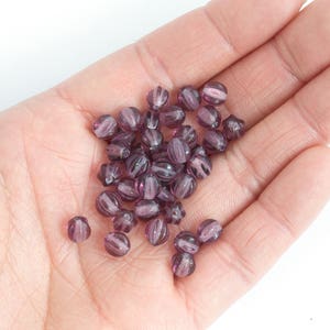 May include: A handful of translucent, purple glass beads. Each bead is oval-shaped with vertical ridges and a lighter purple core. The beads are scattered across a light-colored palm.