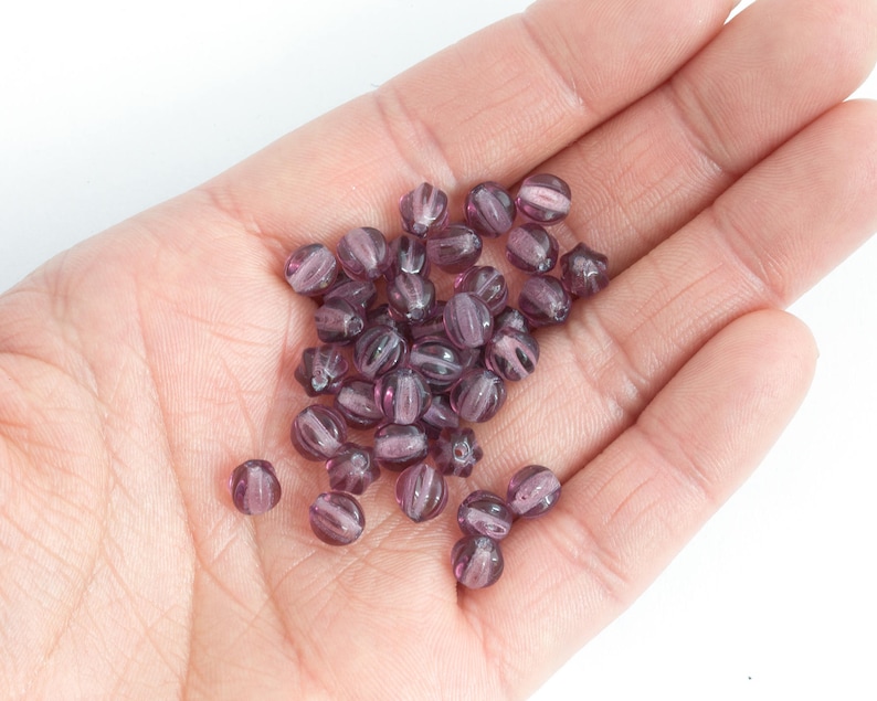 May include: A handful of translucent, purple glass beads. Each bead is oval-shaped with vertical ridges and a lighter purple core. The beads are scattered across a light-colored palm.