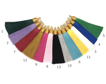 May include: A collection of thirteen colorful tassels, each with a gold-colored cap and loop. The tassels are arranged in a semi-circle, showcasing a variety of colors including green, purple, red, gold, pink, black, gray, blue, yellow, and white.