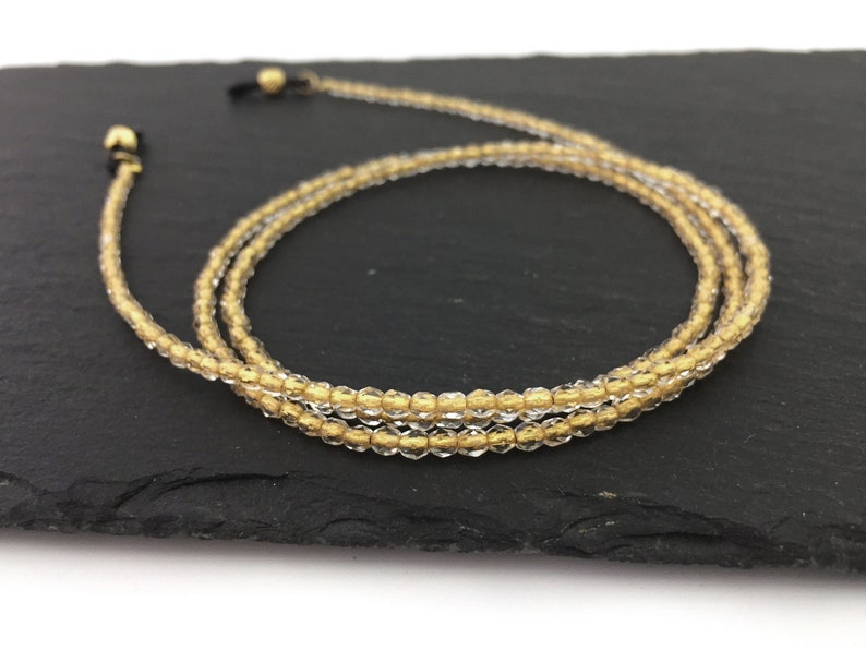 May include: A gold and clear beaded eyeglass chain with gold-toned metal clasps. The chain is coiled on a black surface.