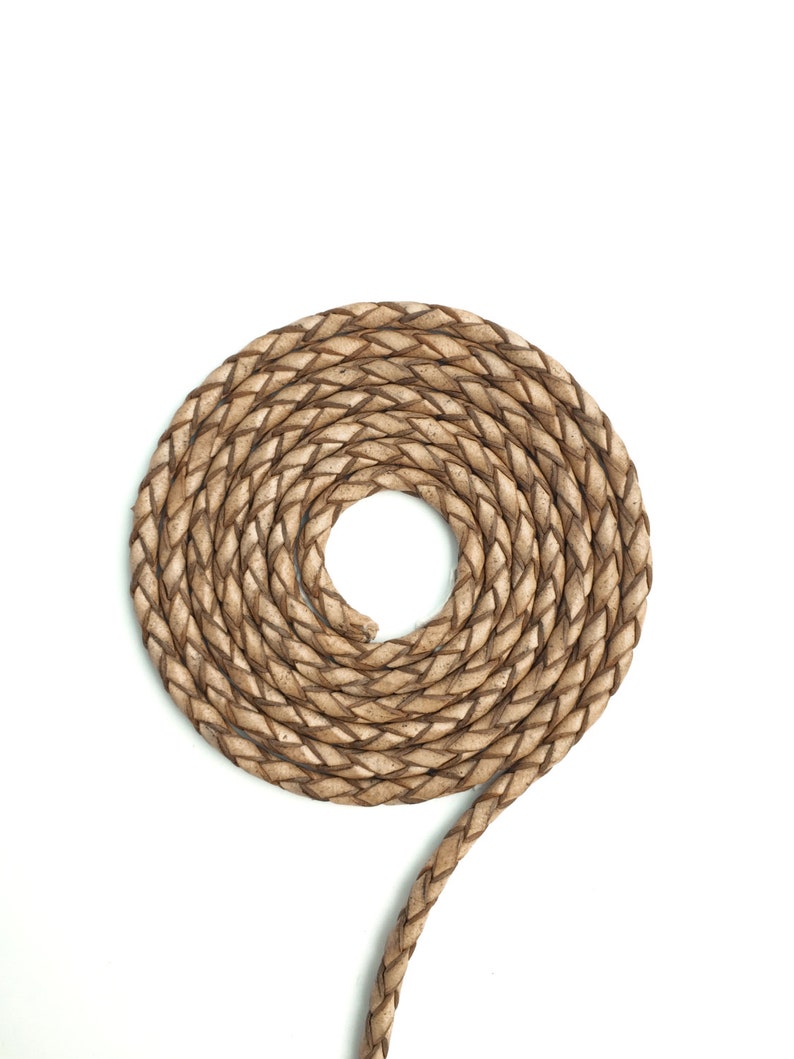 May include: Coiled, light brown braided leather cord against a white background. The cord is tightly woven, creating a circular pattern with a loose end extending outward. The texture of the leather is visible, showing variations in color and tone.