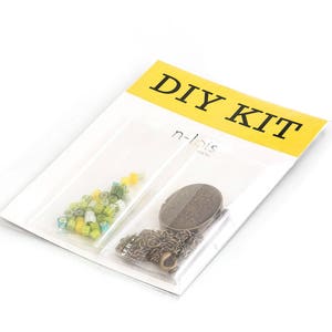 Op de afbeelding: DIY-kit met een geel en zwart label met de tekst "DIY KIT". De kit bevat een klein zakje met groene en gele kralen, een metalen ketting en een ronde metalen hanger.