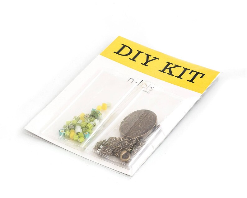Op de afbeelding: DIY-kit met een geel en zwart label met de tekst "DIY KIT". De kit bevat een klein zakje met groene en gele kralen, een metalen ketting en een ronde metalen hanger.