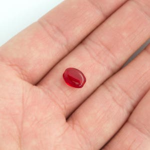 May include: A single, oval-shaped, red gemstone. The gemstone is smooth and shiny.