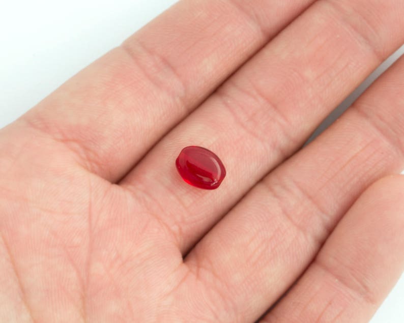 May include: A single, oval-shaped, red gemstone. The gemstone is smooth and shiny.