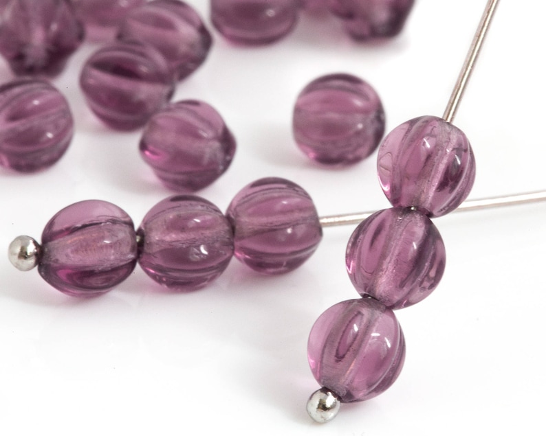 May include: Close-up of several translucent, purple glass beads with a ribbed texture. Some beads are strung on a silver wire, while others are scattered. The beads have a spherical shape and a glossy finish.