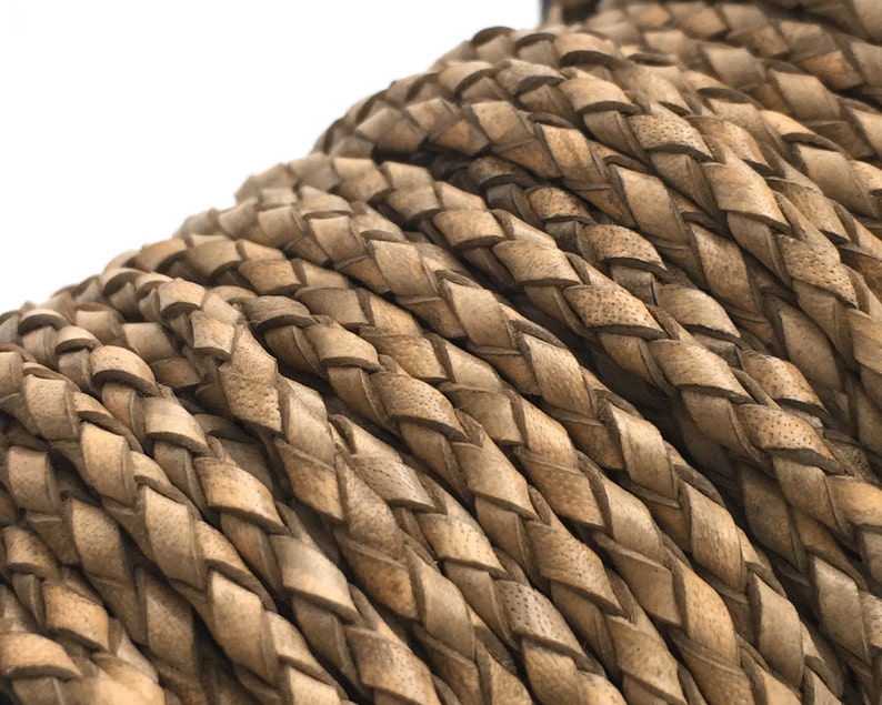 May include: Close-up of a coil of braided leather cord. The cord is a light brown color with a textured surface. The braid is tightly woven, creating a pattern of diagonal lines. This cord is suitable for jewelry making and crafts.