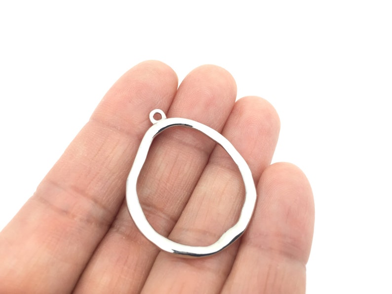 May include: A silver-toned metal pendant charm featuring an irregular, organic, open-frame shape with a smooth, reflective surface. The charm includes a small, circular jump ring at the top for attachment, showcasing a simple, hollow design.