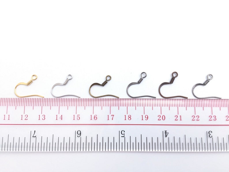 May include: Five earring hooks in different finishes: gold, silver, bronze, black, and gunmetal. The hooks are lined up on a ruler, with the gold hook at the 12-inch mark, the silver hook at the 14-inch mark, the bronze hook at the 16-inch mark, the black hook at the 18-inch mark, and the gunmetal hook at the 20-inch mark.