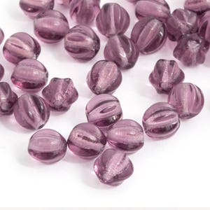 May include: A collection of translucent, purple glass beads. Each bead has a rounded shape with vertical ridges and a swirl pattern. The beads are scattered on a white surface, creating a visually appealing arrangement.