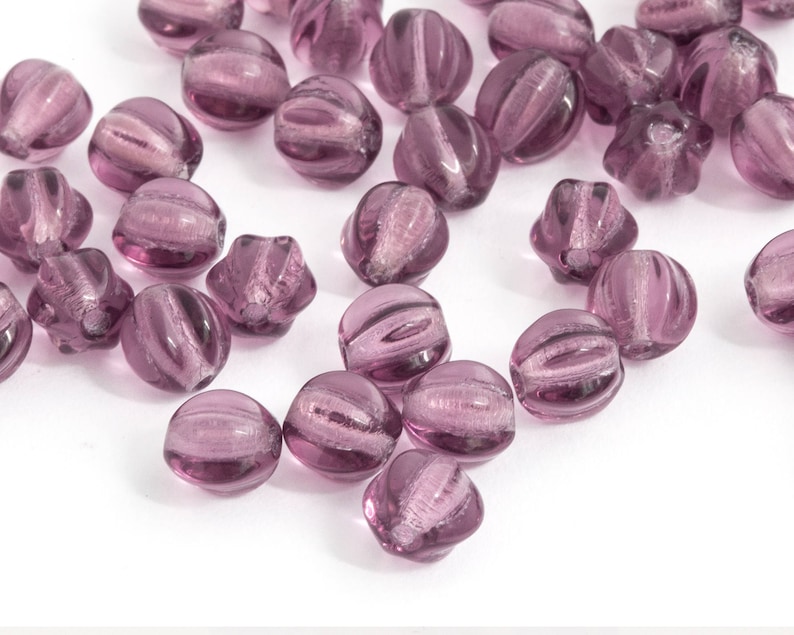 May include: A collection of translucent, purple glass beads. Each bead has a rounded shape with vertical ridges and a swirl pattern. The beads are scattered on a white surface, creating a visually appealing arrangement.