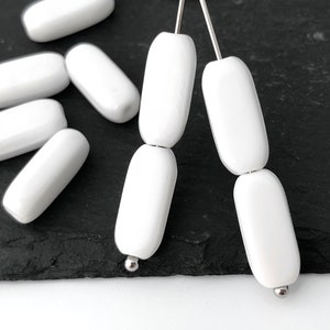 Könnte beinhalten: Weiße Keramikperlen auf einem silbernen Draht gefädelt. Die Perlen sind oval geformt und haben eine glatte, glänzende Oberfläche.