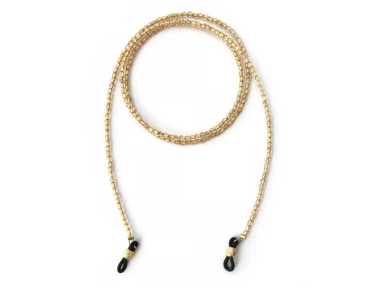 May include: A gold beaded eyeglass chain with black silicone ends. The chain is made of small, round beads and is approximately 28 inches long.