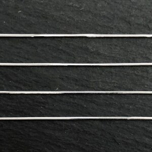May include: Close-up of four silver snake chains against a dark, textured background. The chains are thin and have a smooth, reflective surface, ideal for jewelry making or crafting. The chains are parallel to each other.