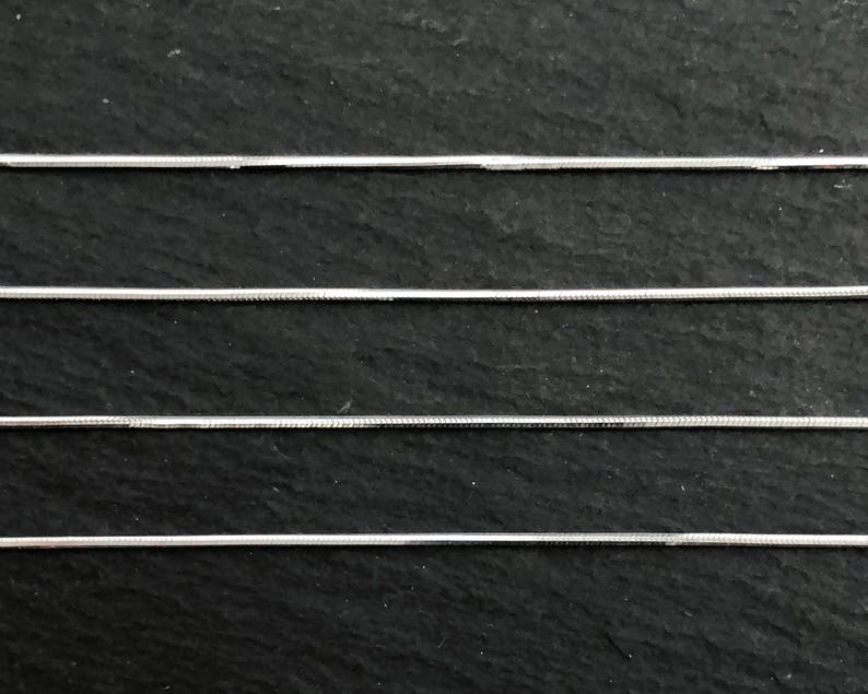 May include: Close-up of four silver snake chains against a dark, textured background. The chains are thin and have a smooth, reflective surface, ideal for jewelry making or crafting. The chains are parallel to each other.