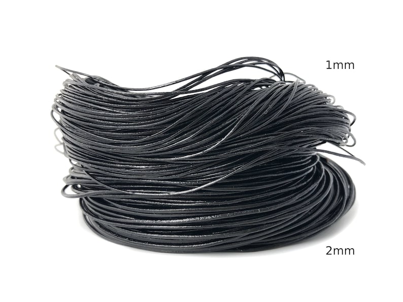 Puede incluir: Una bobina de cord&oacute;n negro, probablemente de cuero o un material similar. La imagen muestra el grosor del cord&oacute;n, con etiquetas que indican di&aacute;metros de 1 mm y 2 mm. El cord&oacute;n est&aacute; sobre un fondo blanco.
